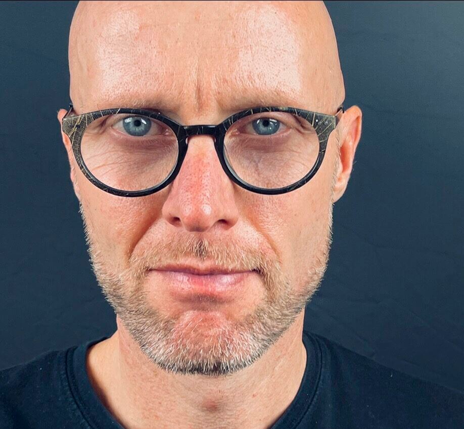 Professional trainer and coach wearing glasses, looking confidently at the camera against a dark background.